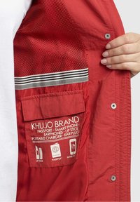 Red jacket interior featuring a mesh panel, labeled pocket for smartphone and accessories, and a striped trim along the edge.