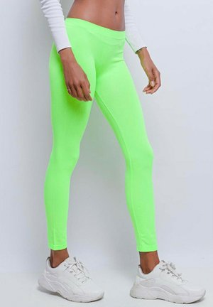 Personne portant des leggings verts fluo vifs, un haut blanc court à manches longues et des baskets blanches épaisses, debout devant un fond uni.