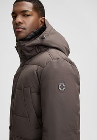 Giubbotto imbottito in marrone scuro con colletto alto e fodera in pelliccia. Caratterizzato da una tasca sul petto e un logo nero sulla manica sinistra.