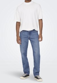 Camiseta blanca de algodón, de corte holgado, combinada con jeans de mezclilla azules, de pierna recta, de tiro medio, y zapatillas negras con cordones blancos.