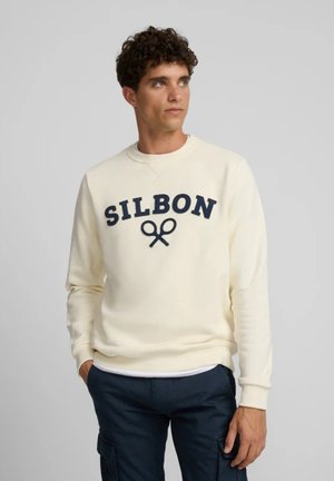 Joven con cabello rizado que lleva una sudadera Silbon color crema y pantalones cargo azul marino, de pie con las manos en los bolsillos.