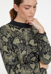 Strickpullover in olivgrün mit einem schwarzen Schlangenprintmuster; verfügt über einen strukturierten schwarzen Rundhalsausschnitt und lockere Ärmel mit gerippten Bündchen.