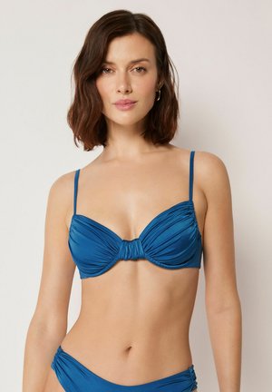 Kvinde iført en blå bikini med rynkede detaljer og tynde stropper, stående mod en ensartet lys baggrund og med fronten mod betrakteren.