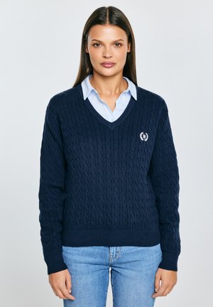 Námořnicky modrý svetr s copánkovým vzorem a výstřihem do V, s erbem na levé straně hrudi a žebrovanými manžetami a lemem. Nositelný přes světle modrou košili.