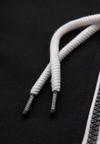 Felpa nera con cappuccio caratterizzata da lacci intrecciati bianchi con terminali neri etichettati "DMBRE" e un particolare dettaglio della cerniera nera texturizzata di lato.