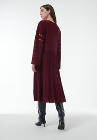 Robe bordeaux à manches longues avec des découpes, longueur genou et silhouette évasée. Associée à des bottes en cuir noir montée jusqu'aux genoux.