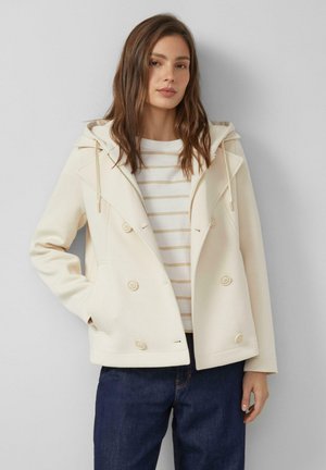 Frau trägt cremefarbene, doppelt geknöpfte Jacke mit Kapuze, gestreiftes weiß-beiges Hemd und dunkelblaue Jeans, steht vor hellgrauem Hintergrund.