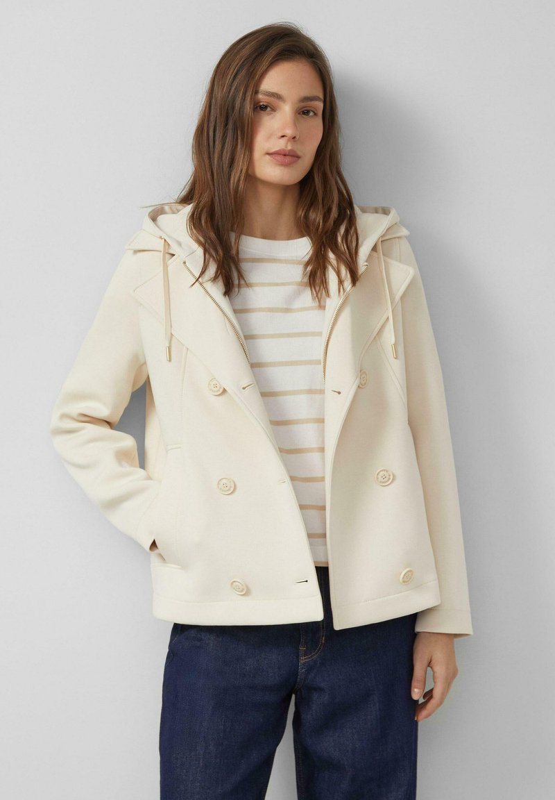 Frau trägt cremefarbene, doppelt geknöpfte Jacke mit Kapuze, gestreiftes weiß-beiges Hemd und dunkelblaue Jeans, steht vor hellgrauem Hintergrund.