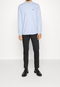 Lichtblauw overhemd met knopen en een kraag, één borstzak en Lacoste-logo, gecombineerd met zwarte broek en zwarte schoenen.