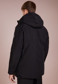 Czarna kurtka puchowa z kapturem, o gładkiej, miękkiej fakturze i naszywce z logo na lewym rękawie. Prosty, nowoczesny design o luźnym dopasowaniu.