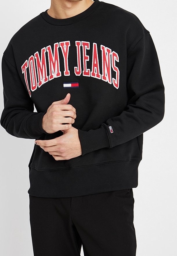 Homme portant un sweat-shirt noir avec "TOMMY JEANS" en lettres rouges et blanches et un petit logo de drapeau sur la poitrine et la manche, les mains jointes devant.