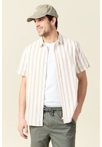 Camisa de manga corta, con botones, de rayas verticales beige y blancas, hecha de tela ligera. Llevada sobre una camiseta blanca y combinada con pantalones de chándal verdes.