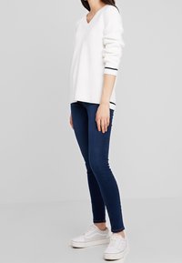 Pull blanc côtelé avec un col en V, manches longues et accents à rayures bleu marine. Associé à un jean slim bleu foncé et des baskets blanches.
