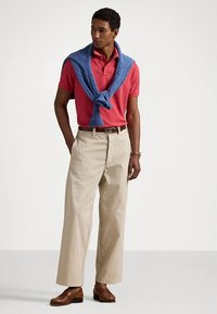 Červené polo tričko, modrý pletený svetr přehozený přes ramena, béžové široké kalhoty, hnědé kožené boty, na levém zápěstí hodinky.