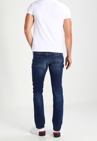Jeans de mezclilla azul con corte slim, cintura media y dos bolsillos traseros, combinados con una camiseta blanca sencilla y zapatillas blancas.