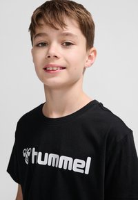 Tricou din bumbac negru cu mâneci scurte, cu un logo alb „hummel” proeminent pe față. Design simplu, guler rotund.