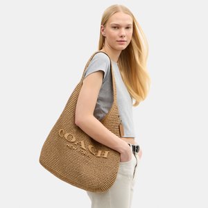 Plavuša žena u sivoj majici i bijelim hlačama drži veliku smeđu tkanu torbu Coach na ramenu.