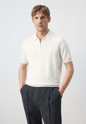 Hombre con polo blanco de manga corta con cremallera y pantalones negros plisados con las manos en los bolsillos, sobre un fondo liso.