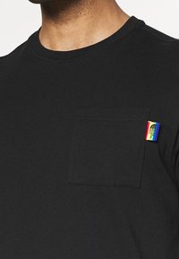 Black cotton t-shirt with a round neckline, featuring a chest pocket and a small multicolored tag on the side.