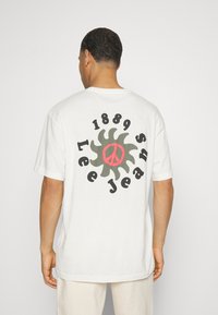 Camiseta blanca de algodón con un gran gráfico en la parte posterior, que incluye un sol estilizado, un símbolo de paz y el texto "1889 Lee Jeans."
