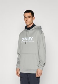 Šviesiai pilka liemenė pagaminta iš minkštos medžiagos su dideliu baltu „OAKLEY“ logotipu. Yra su raišteliu užtraukiama kapuce ir kengūros kišenė.