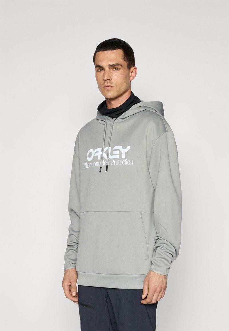 Šviesiai pilka liemenė pagaminta iš minkštos medžiagos su dideliu baltu „OAKLEY“ logotipu. Yra su raišteliu užtraukiama kapuce ir kengūros kišenė.