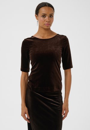 Femme aux cheveux foncés attachés en arrière, portant un haut court en velours marron foncé et une jupe assortie en velours avec un détail froncé.