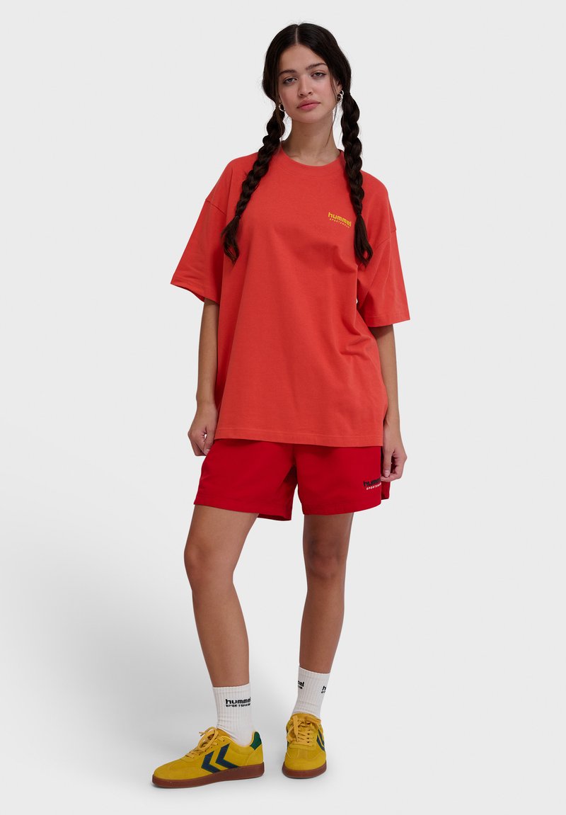 Oranžové oversized tričko so žltým logom, v kombinácii s červenými šortkami, bielymi ponožkami a žltými teniskami so zelenými akcentmi.