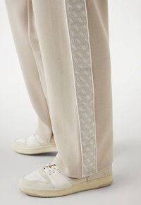Beige Jogginghosen mit einem gemusterten Seitenstreifen, kombiniert mit hellen Sneakern, die eine glatte Textur und subtile Akzente aufweisen.