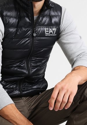 Homme portant une veste matelassée noire avec le logo EA7 Emporio Armani sur un t-shirt à manches longues gris clair, assis avec un bras reposant sur le genou.