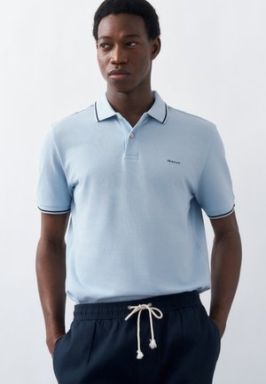 Νέος άνδρας που φοράει ανοιχτό μπλε πουκάμισο πόλο με μαύρες λεπτομέρειες και σκούρο μπλε παντελόνι με κορδόνι, στέκεται με τα χέρια στις τσέπες μπροστά σε λευκό φόντο.