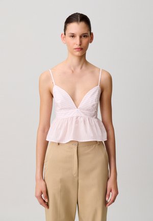 Jeune femme aux cheveux attachés, portant un haut sans manches à rayures roses et un pantalon beige taille haute, sur un fond clair uni.