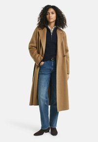 Brauner Wollmantel mit großen Revers über einem marineblauen Zopfmuster-Pullover, kombiniert mit blauen Jeans und dunkelbraunen Stiefeletten.