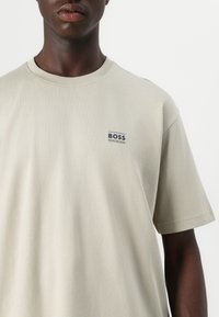 Beige bavlněné tričko s krátkým rukávem, kulatým výstřihem a malým modrým logem "BOSS" na levé hrudi. Měkká textura, klasický střih.