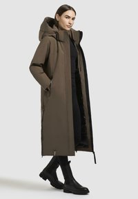 Parka lunga e marrone, impermeabile, con cappuccio, chiusura frontale con zip e tasche laterali. Interno nero, indossata sopra un completo nero con stivali neri.