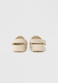 Ανοιχτό μπεζ Crocs με κλειστή φτέρνα, δύο λεπτές λουρίδες, υφασμένο πέλμα και σόλα με σχέδιο. Το όνομα της μάρκας "crocs" είναι ορατό στα λουριά.