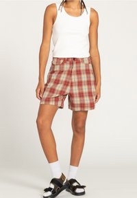 Červené kostkované šortky se žlutými a modrými akcenty, s knoflíkovým zapínáním a kapsami. Kombinovány s bílým tílkem a černými sandály.
