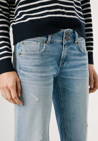 Gaiši zili denimpantaloni ar nelielu izbalēšanu, kas aprīkoti ar vairākiem kabatas, nolietojuma detaļām un divu pogu aizdari jostasvietā.