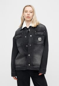 Ξανθιά γυναίκα φορώντας υπερμεγέθη ξεθωριασμένο μαύρο τζιν μπουφάν με κουμπιά μπροστά, λευκό πουκάμισο με γιακά και μαύρη γραβάτα, στέκεται μπροστά σε απλό φόντο.