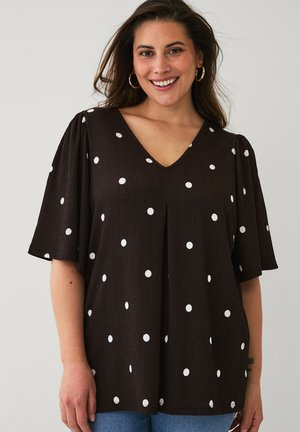 Kobieta uśmiechająca się, ubrana w czarną bluzkę z dekoltem w serek, białymi grochami i krótkimi, zwiewnymi rękawami na jednolitym tle.