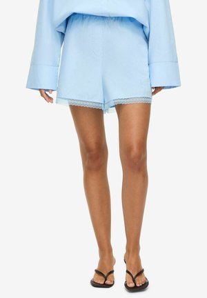 Femme portant un short ample bleu clair avec un ourlet en dentelle et un top assorti, associé à des sandales noires à brides, debout sur fond blanc.