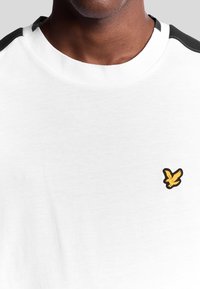 Vit kortärmad t-shirt i mjukt material, med en svart axeldetalj och en liten gul och svart logotyp på bröstet.