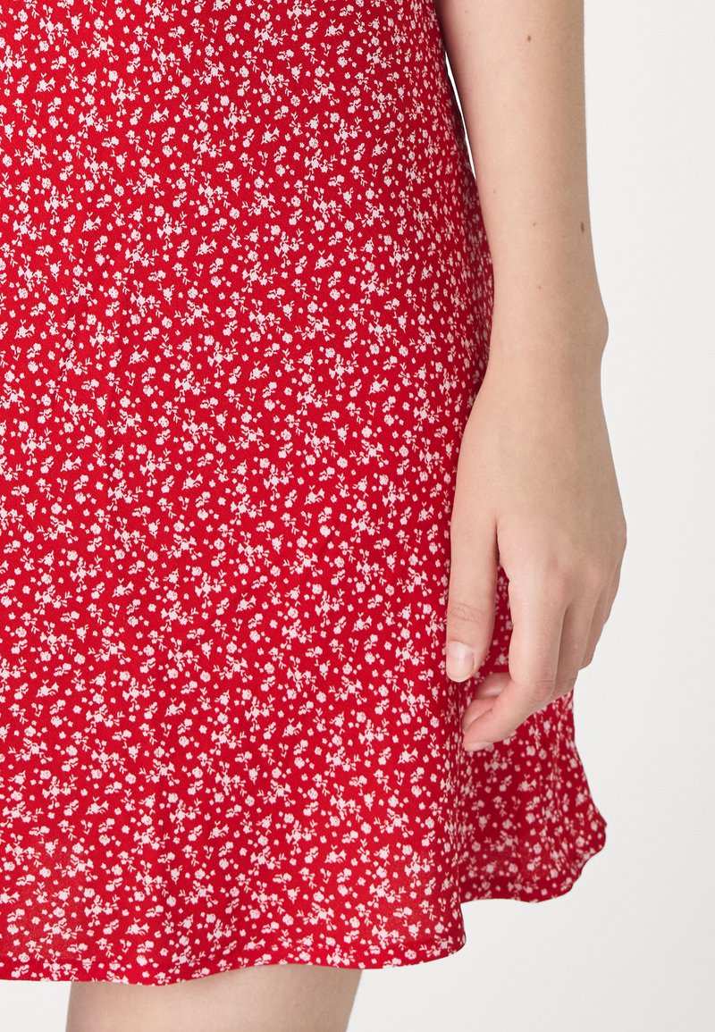 Rotes Blumenkleid aus leichtem Stoff mit kleinen weißen Blumen, einem ausgestellten Saum und einer glatten Textur. Eine Hand ruht auf dem Kleid.