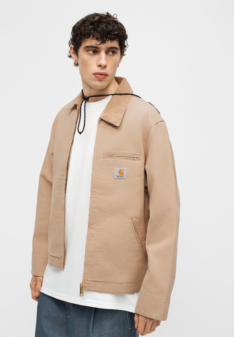 Giovane uomo che indossa una giacca beige con zip sopra una maglietta bianca e jeans blu, che guarda verso sinistra su uno sfondo neutro.