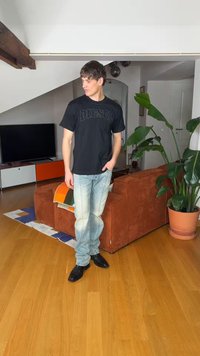 Jonge man draagt een zwart Diesel-shirt en versleten spijkerbroek, staat in een woonkamer met houten vloeren, een oranjebruine bank en grote groene planten.