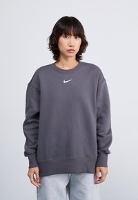 Grijze sweatshirt met een ronde hals, geribde mouwen en zoom, met een witte Nike-logo op de borst. Draagt een lichte spijkerbroek.