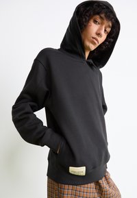 Tânăr cu părul creț, îmbrăcat cu un hanorac negru cu glugă având un eticheta "Studio Edition" și pantaloni maro cu carouri, cu mâinile în buzunare.