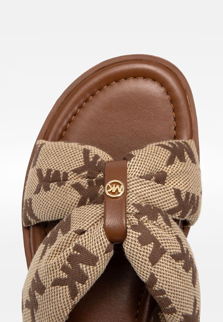 Nahaufnahme einer braunen Ledersandale mit einer beigefarbenen Stoffschleife, die ein dunkelbraunes Muster und ein goldenes rundes Logo in der Mitte aufweist.