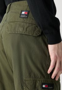 As calças cargo de cor verde-azeitona apresentam vários bolsos, um tecido texturizado e uma etiqueta de marca com cores vermelho, branco e azul na parte de trás.