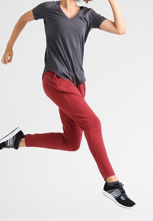 Person i midthop iført en mørkegrå V-hals trøje, røde bukser og sorte sportssko med hvide såler på en ensartet lys baggrund.